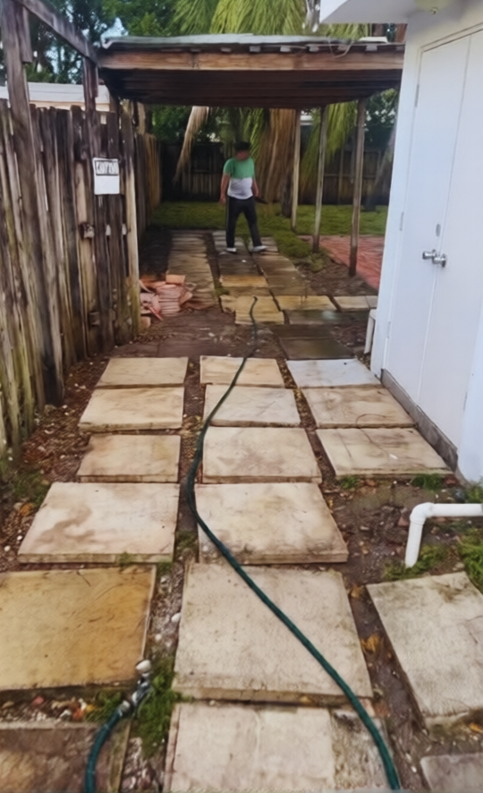 A person stands under a covered patio in a yard with concrete stepping stones and a garden hose extending across the path.