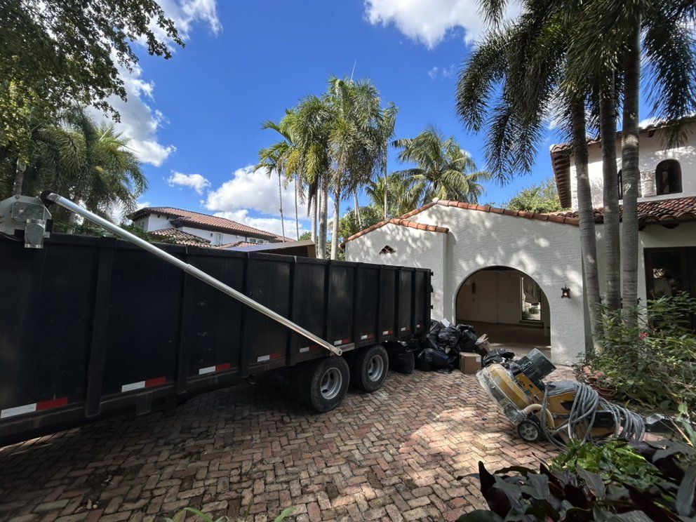 A large black dumpster trailer sits on a paved driveway in front of a white house with palm trees and a blue sky.