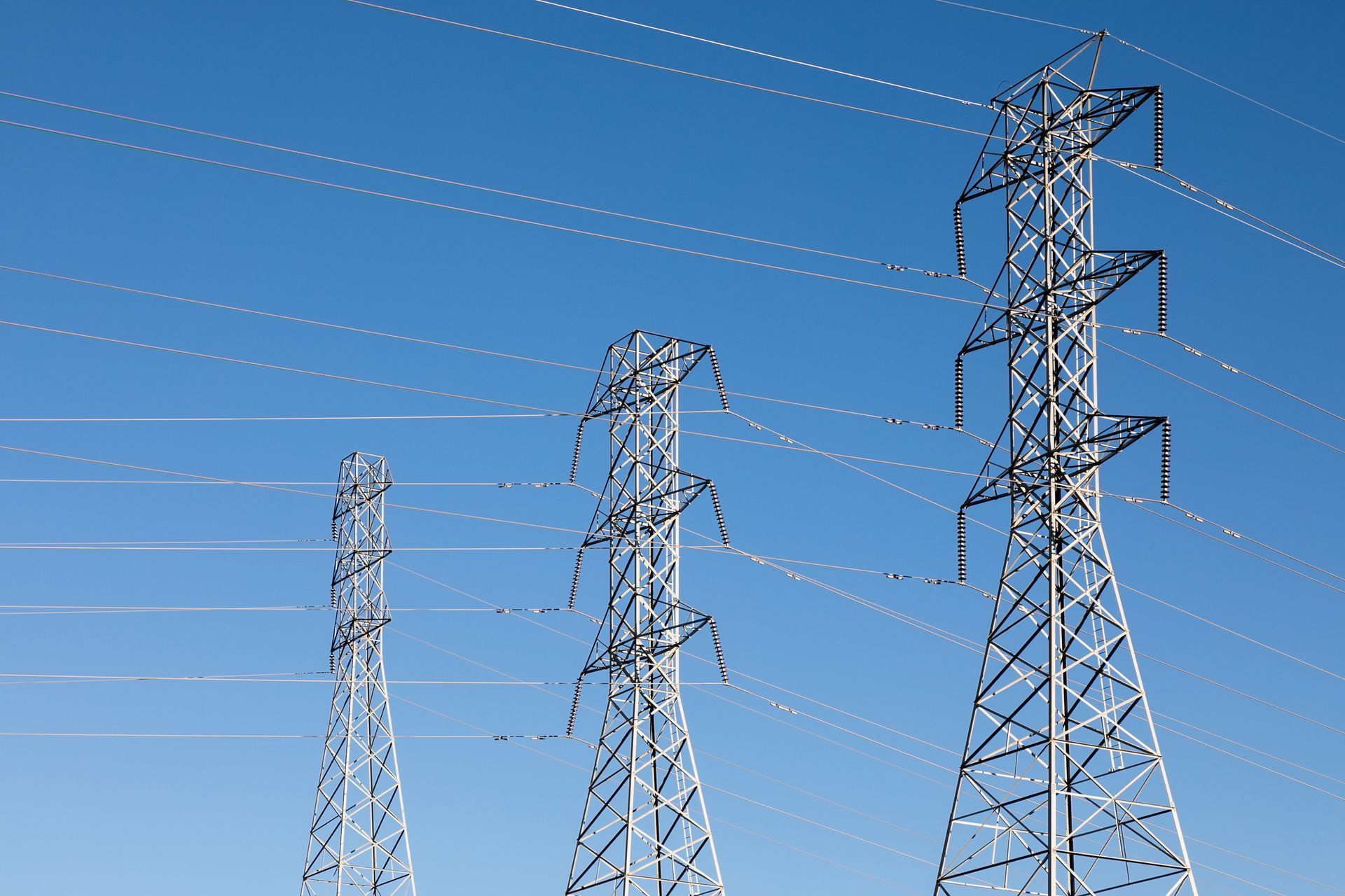 Three metal electricity pylons against a clear blue sky. Overhead power lines stretch across the frame.