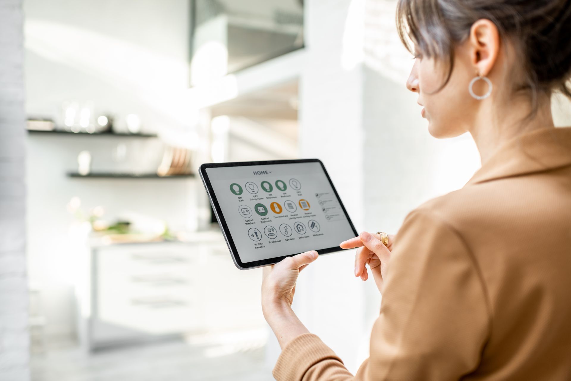 Woman in a tan blazer uses a tablet to control smart home features in a modern kitchen.