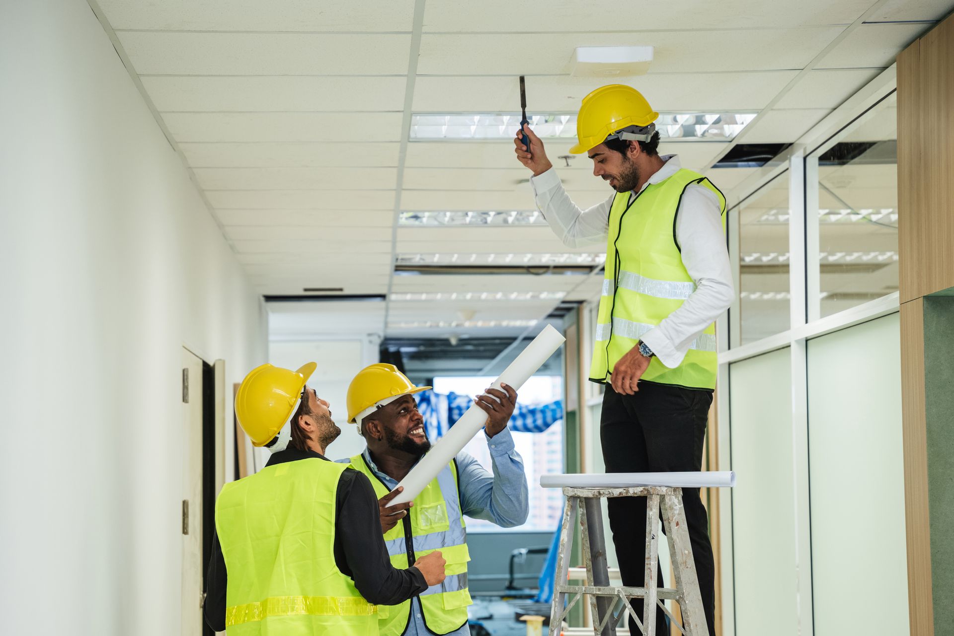 Three construction workers examining ceiling, one on a ladder. They wear yellow helmets and vests.