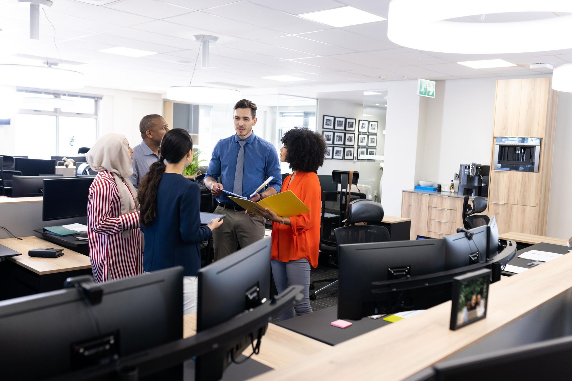 Group of people discussing documents in a brightly lit office, standing near desks with computer monitors.