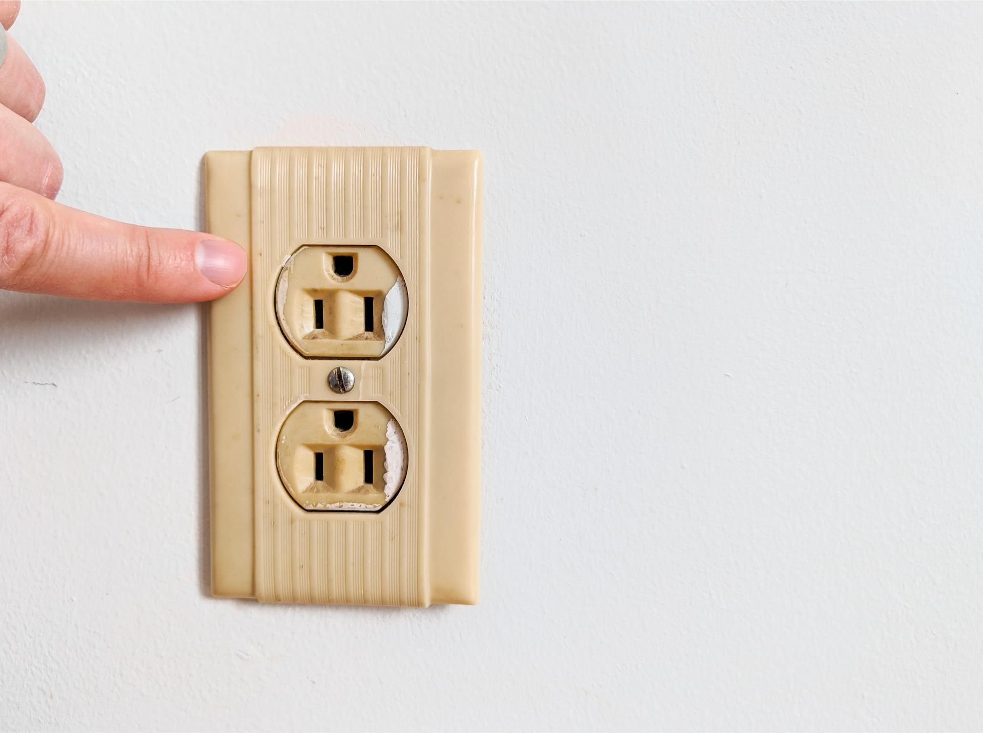 Finger pointing at a beige electrical outlet on a white wall.
