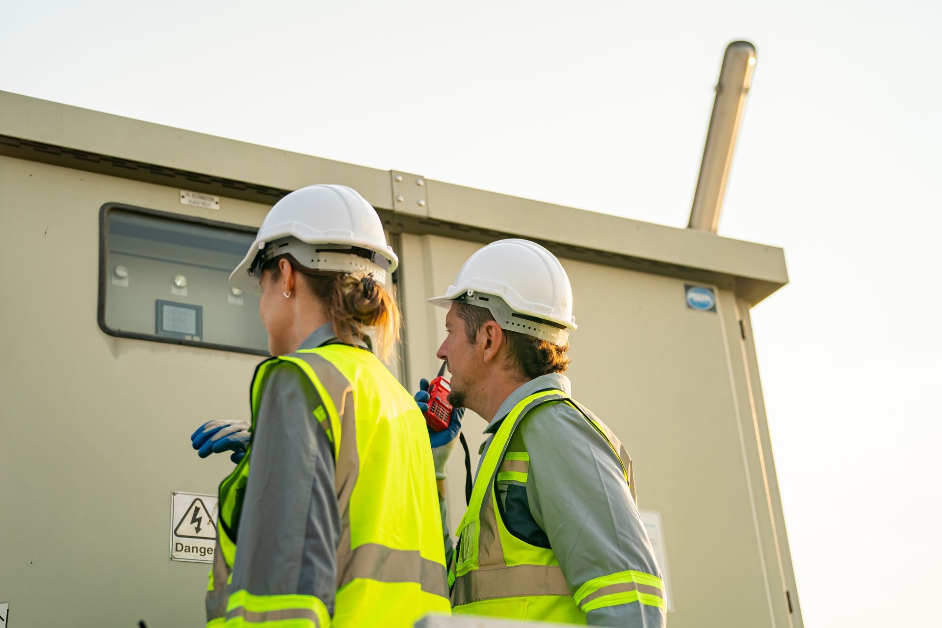 Two people in safety vests and hard hats inspecting electrical equipment outdoors.