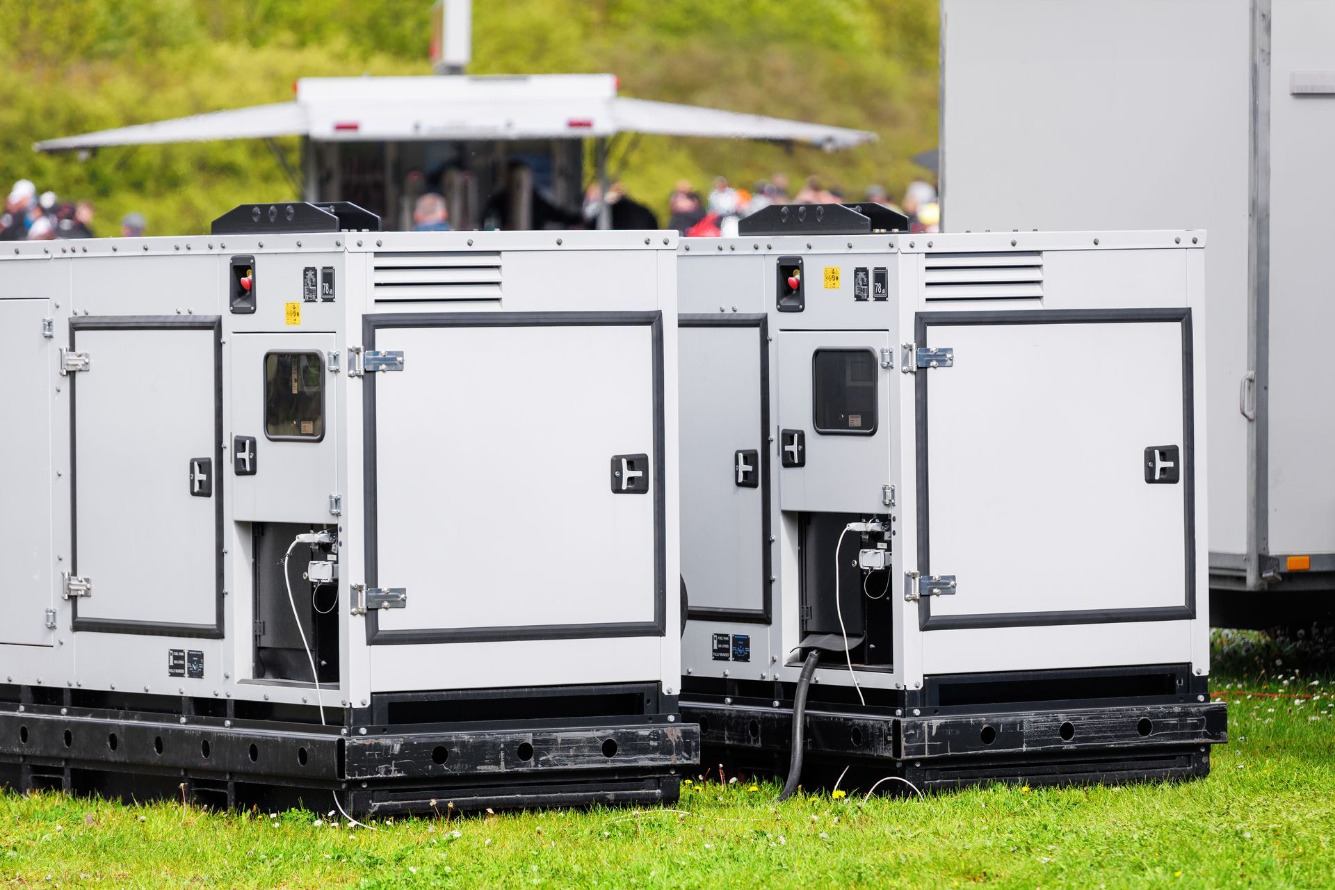 White industrial generator units on wheels parked on grass outdoors, with a crowd blurred in the background