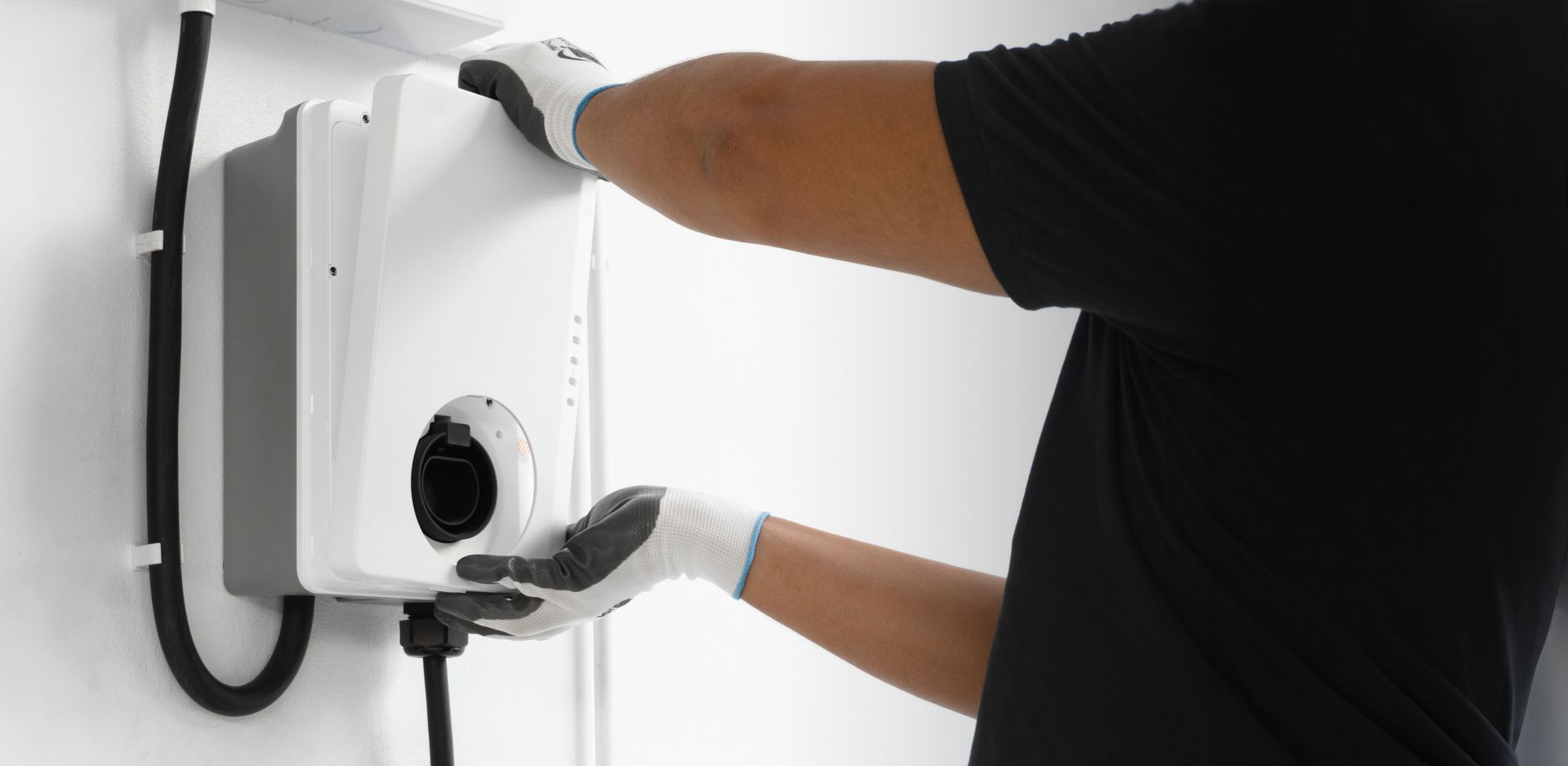 Person adjusting a wall-mounted white device with gloved hands and a black cable attached