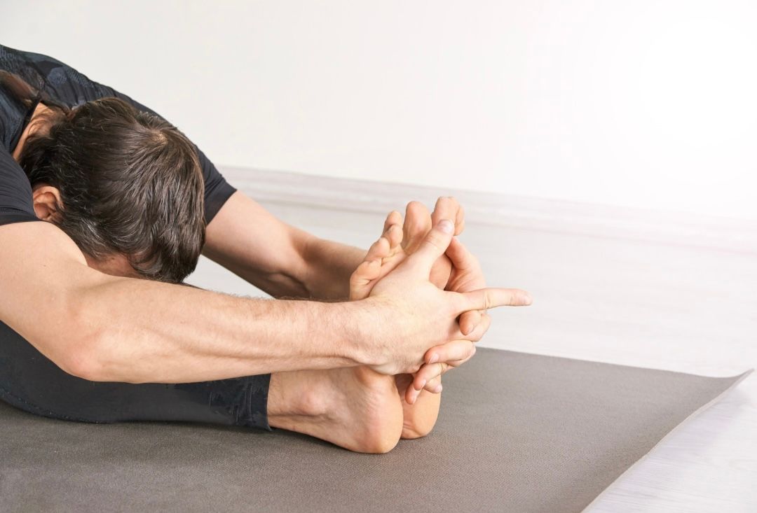 Person performing a seated forward bend, touching feet, on a gray yoga mat.