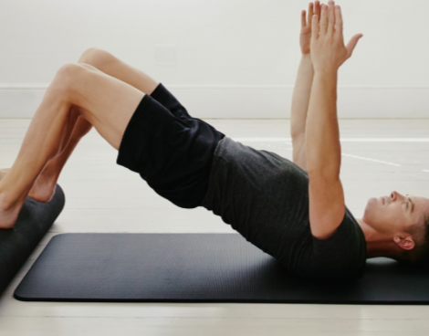 Man doing bridge exercise on mat, hands raised, feet on a roller. Black shorts, grey shirt. Indoors.