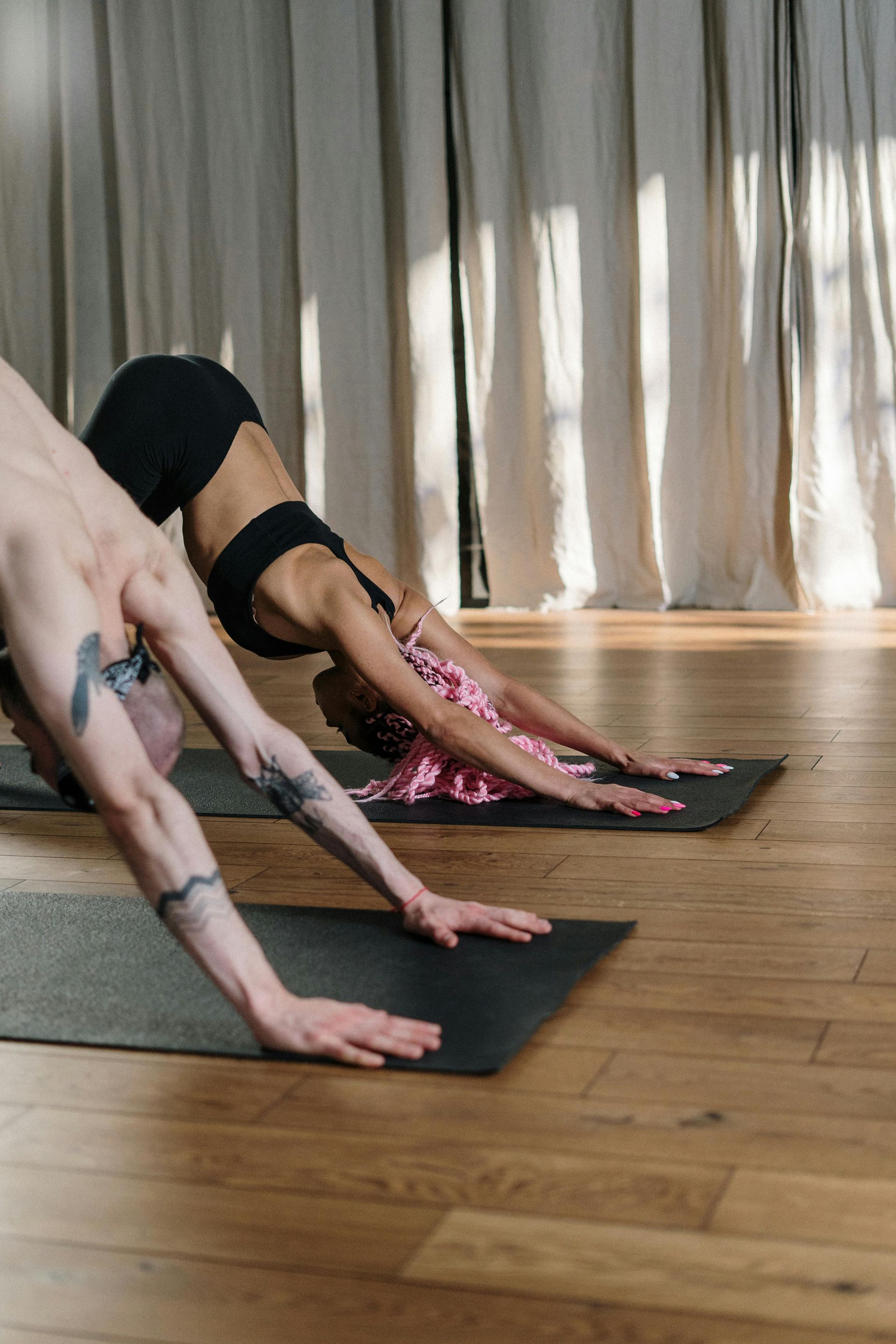 Two people in yoga studio, doing downward-facing dog pose on mats, with a wooden floor and light curtains in background.