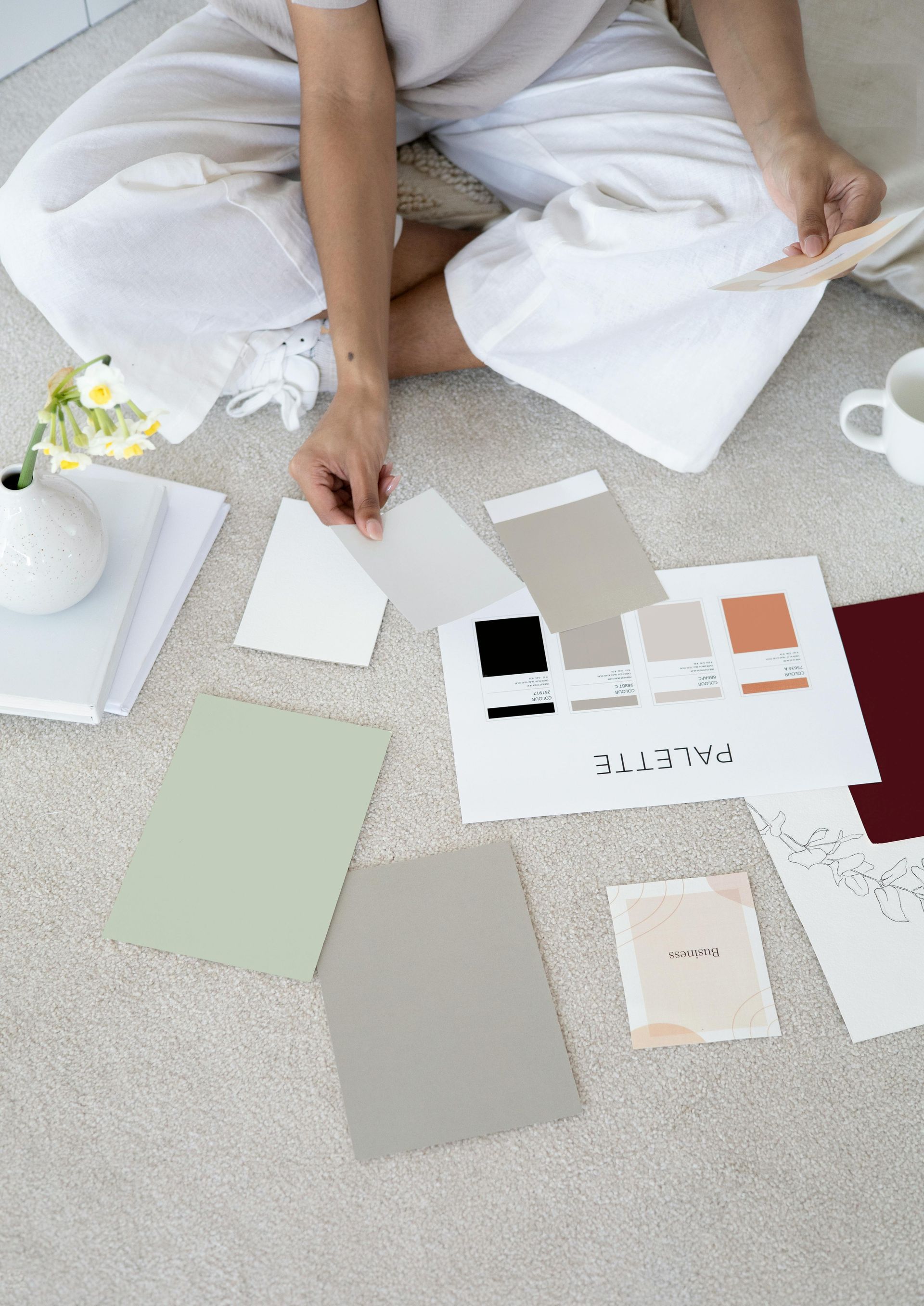 Person seated on floor, reviewing paint swatches, with a notebook, mug, and vase nearby.