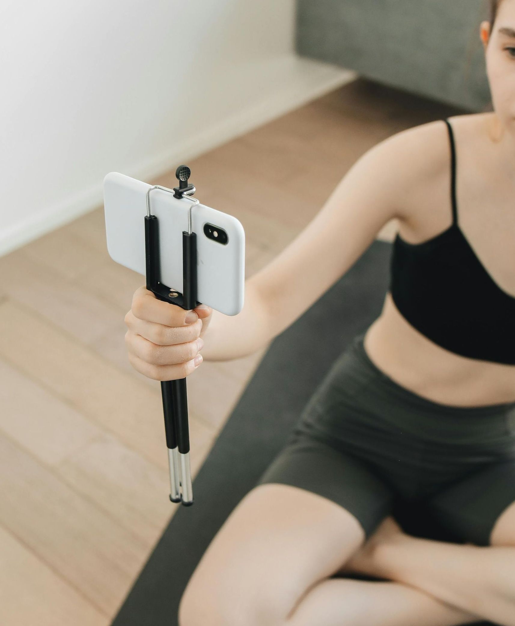 Woman in workout clothes using a phone on a tripod, seated on a yoga mat.