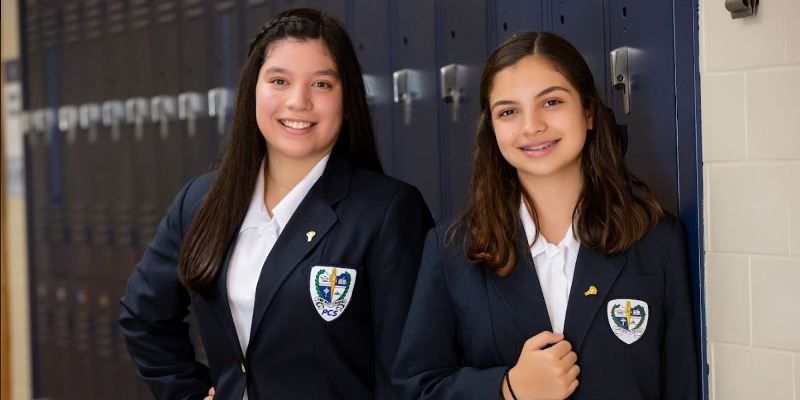 Two girls in school uniforms are standing next to each other in front of lockers.