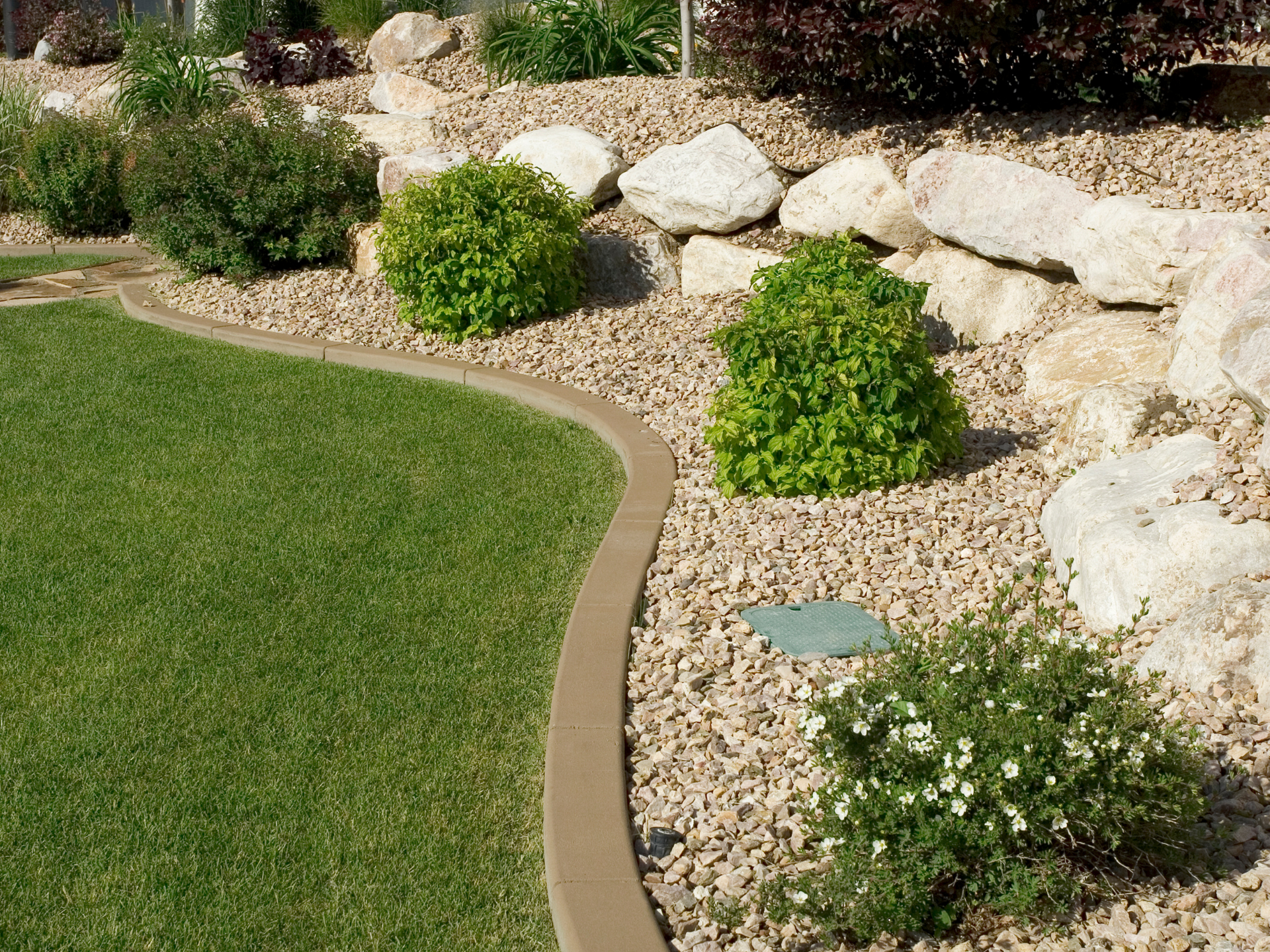 Lush green grass borders a rock garden with bushes, and a retaining wall.
