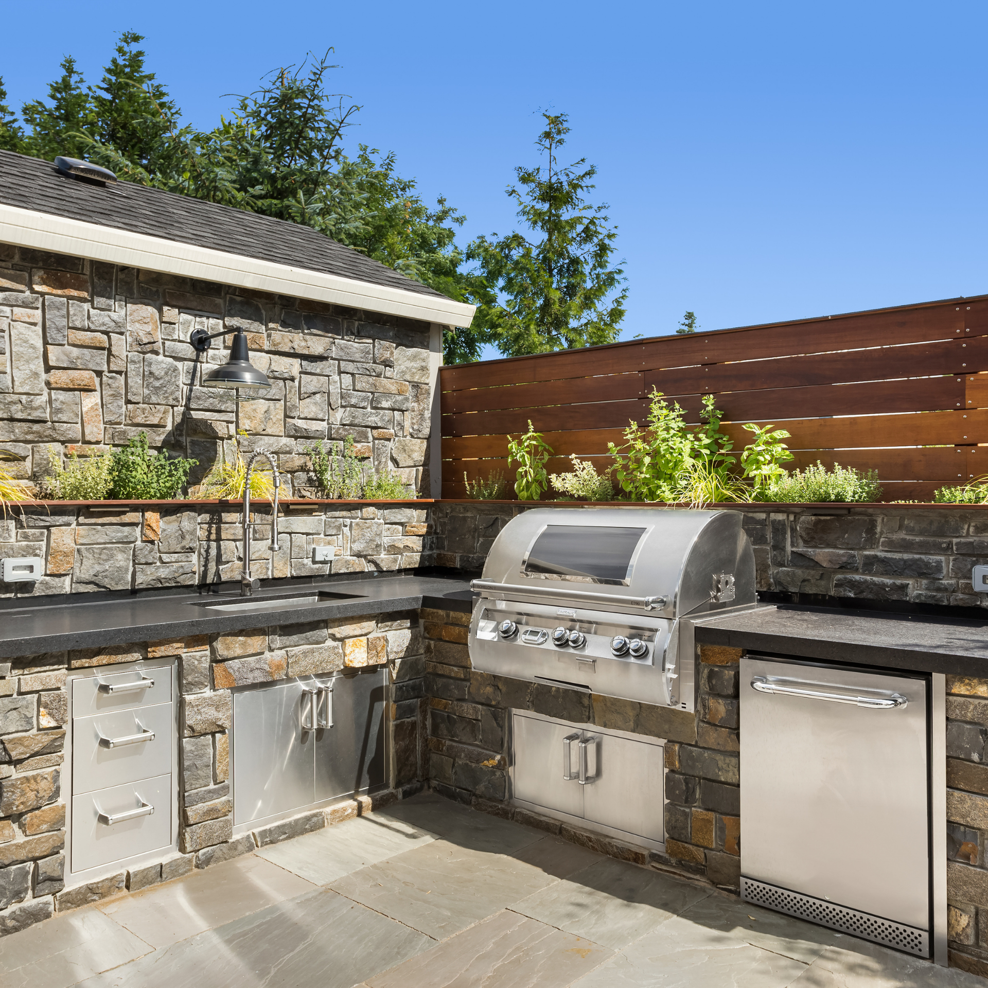 Outdoor kitchen with stone walls, stainless steel grill, cabinets, and a refrigerator.