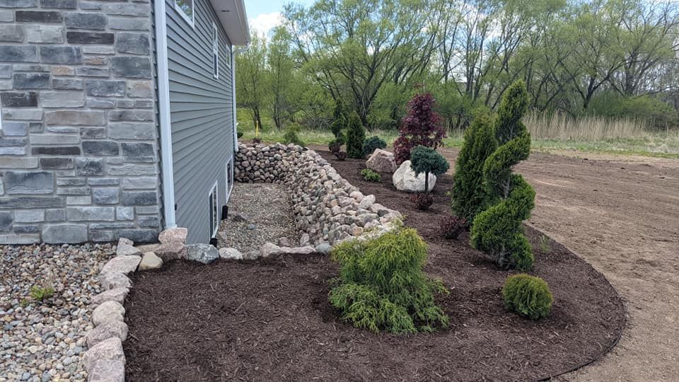 Landscaped garden bed with dark mulch and various green and red shrubs next to a house with stone accents.