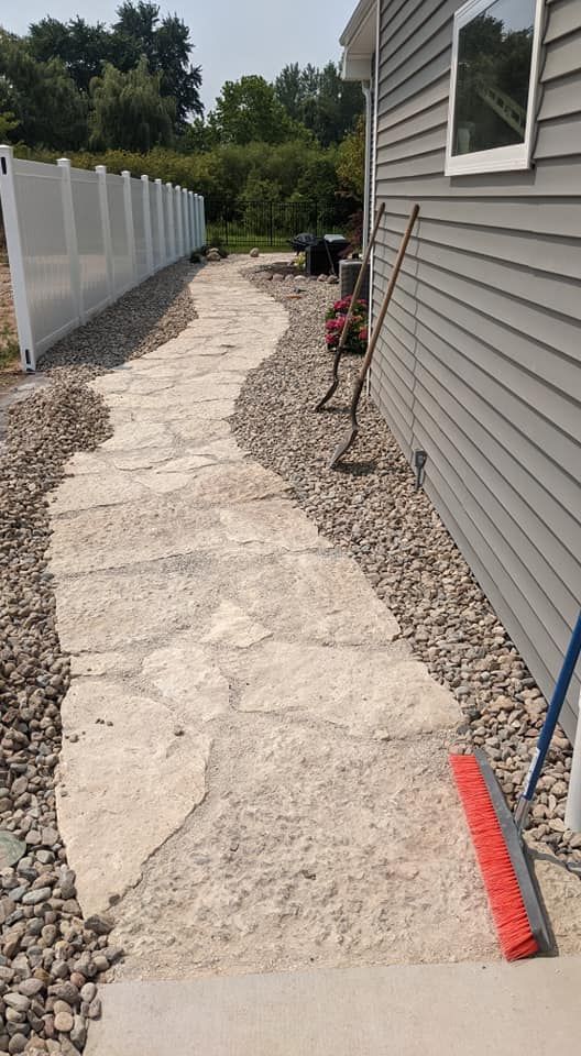 Stone pathway alongside a house with gray siding, a white fence, and a red push broom.