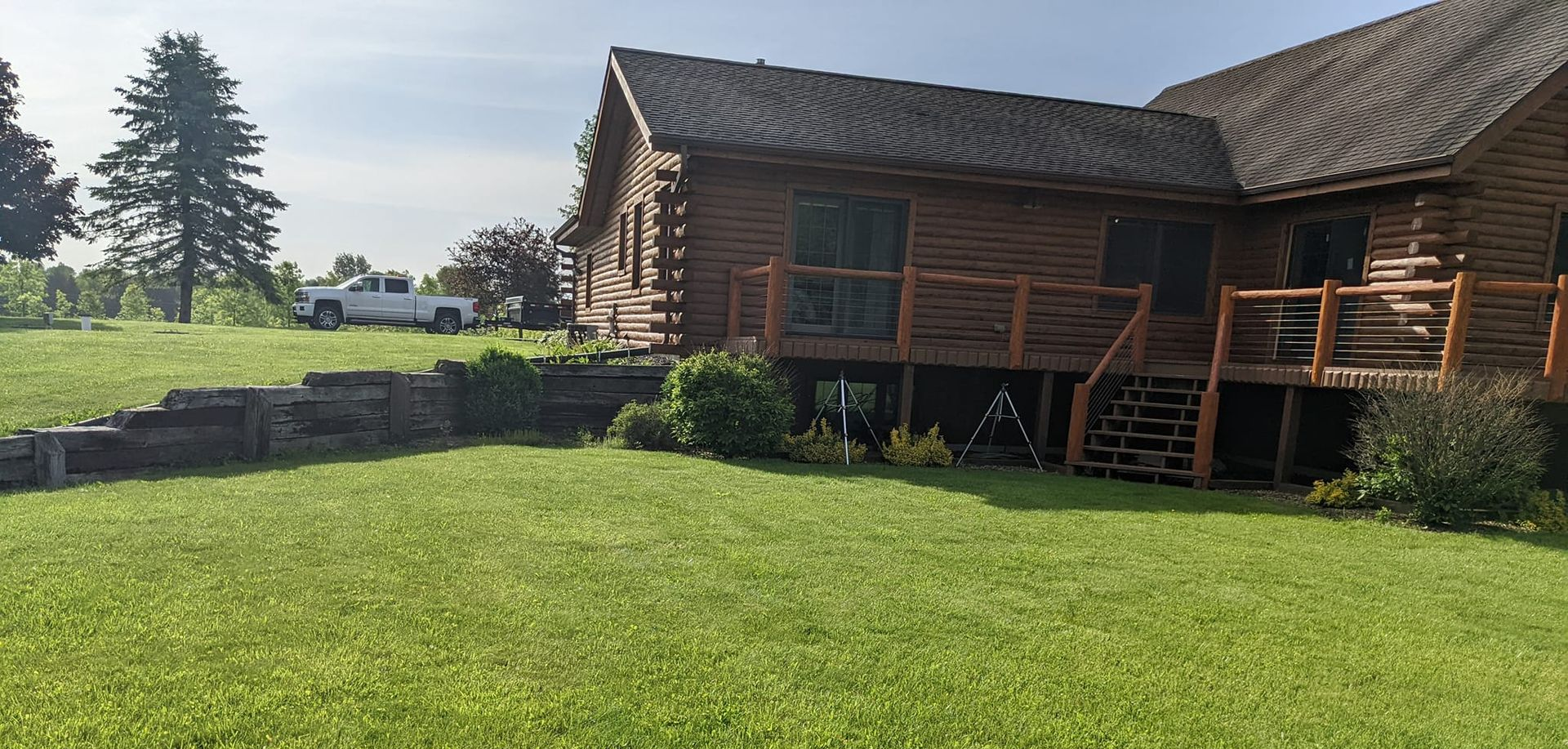 A log cabin with a wooden deck and a grassy yard. A white truck is parked in the distance.