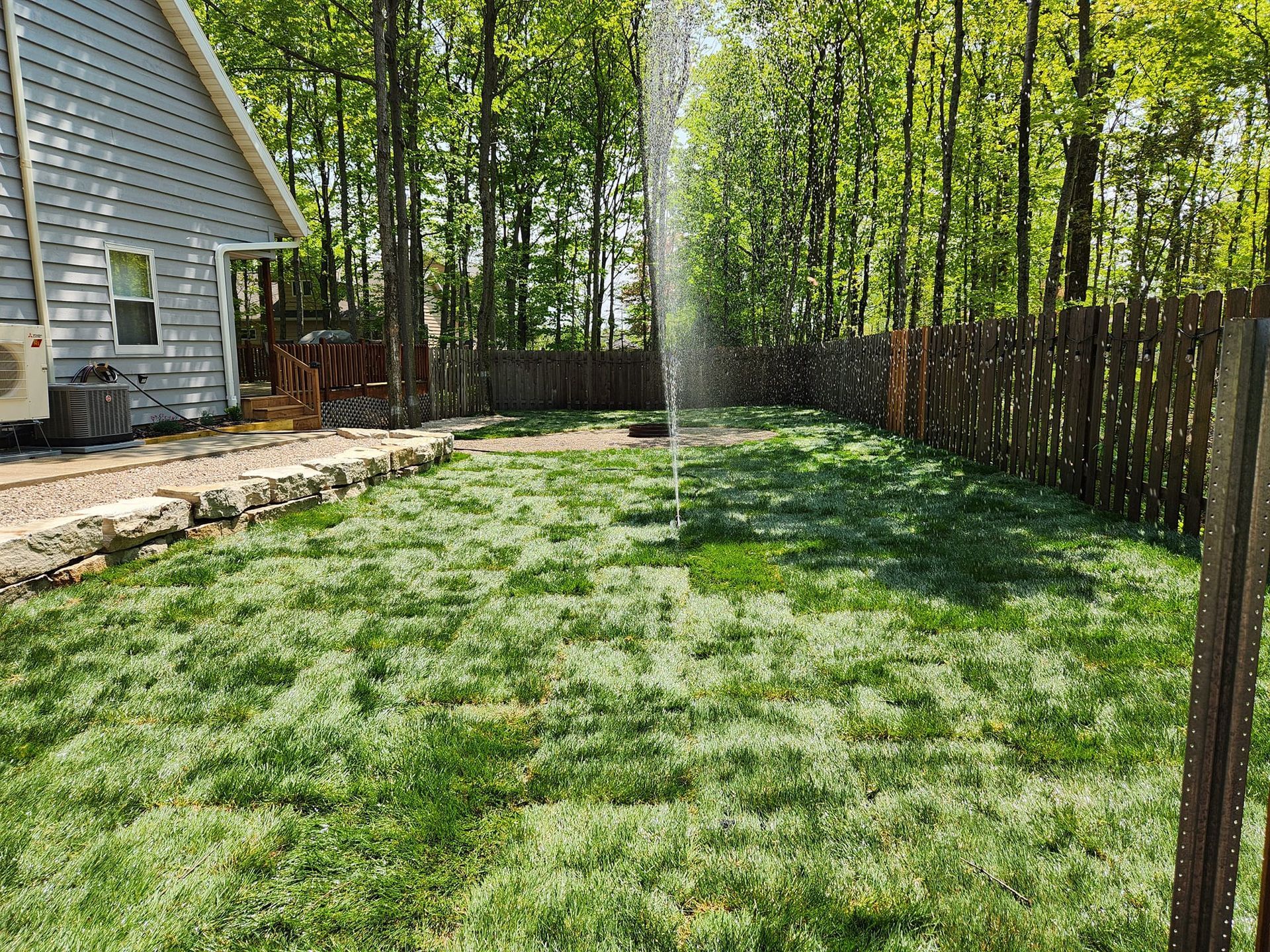 Lawn with sprinkler spraying water, wooden fence, house on the left, trees in background, sunny day.