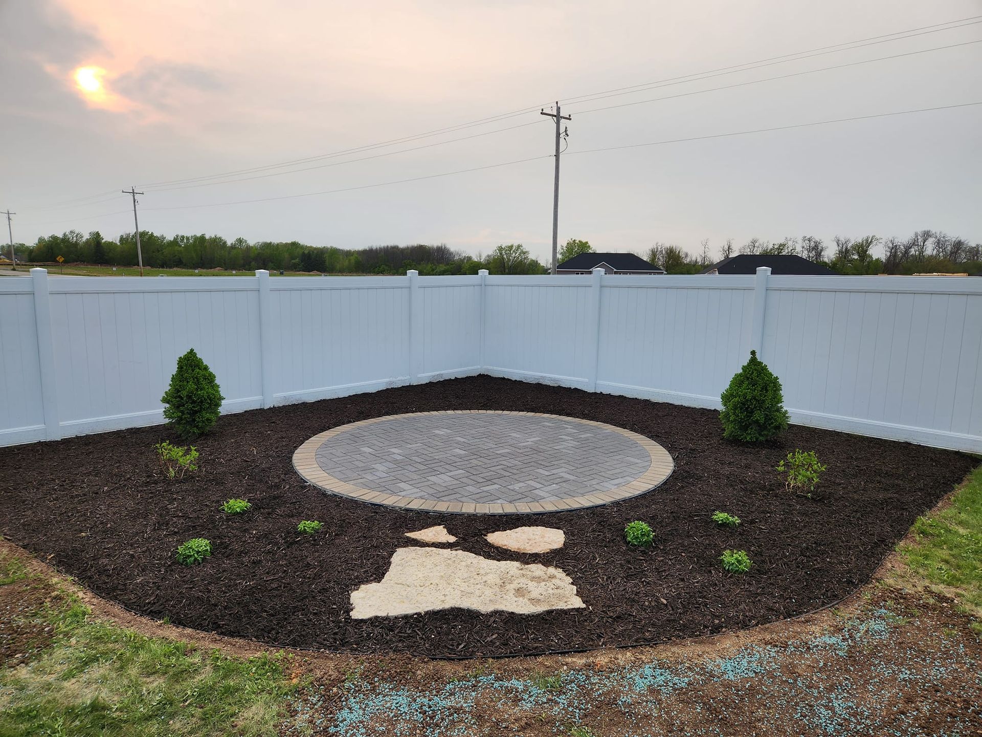 A landscaped garden bed with a stone seating area and two small trees, surrounded by a white fence.