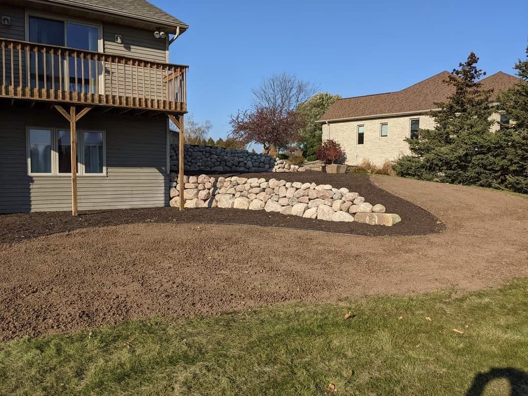 House with wooden deck, rock retaining wall, and brown mulch in the yard. Blue sky background.