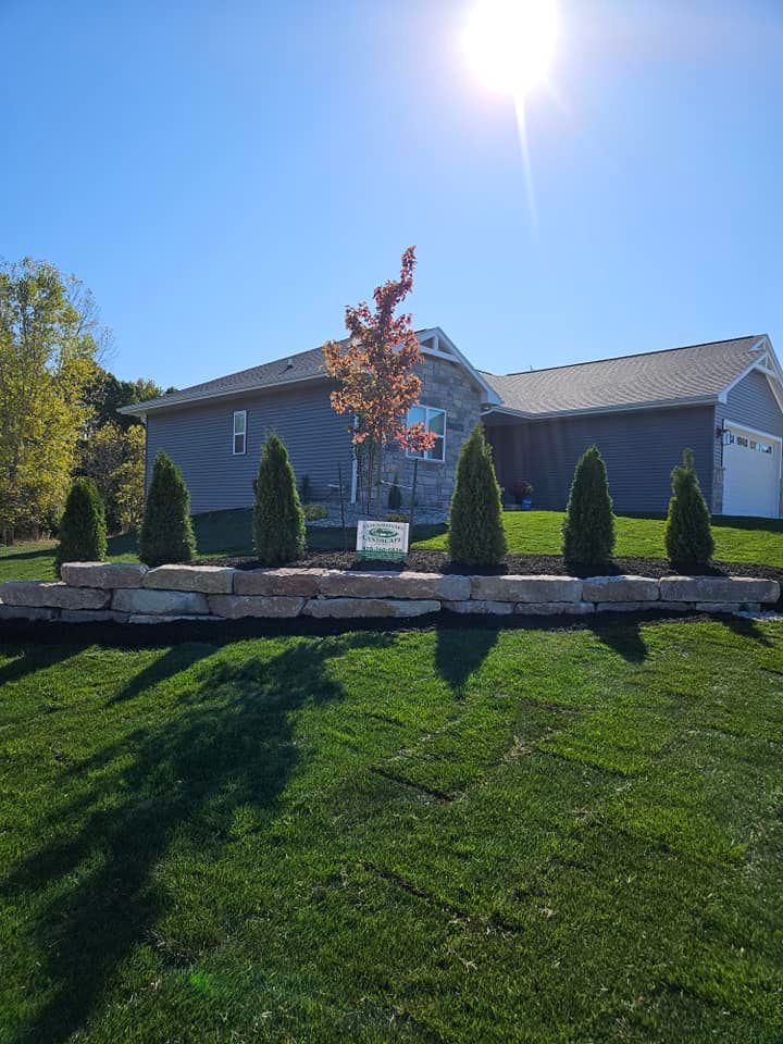 Sunny day view of a house with landscaping; trees, shrubs, stone wall, and green lawn.