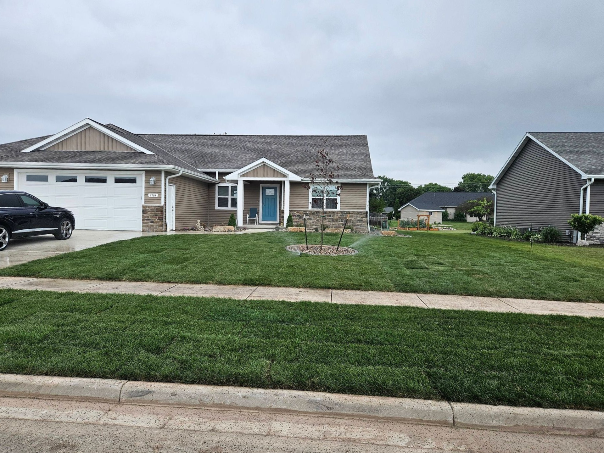 A light brown house with a green lawn. Sprinkler in front yard. Cloudy sky.