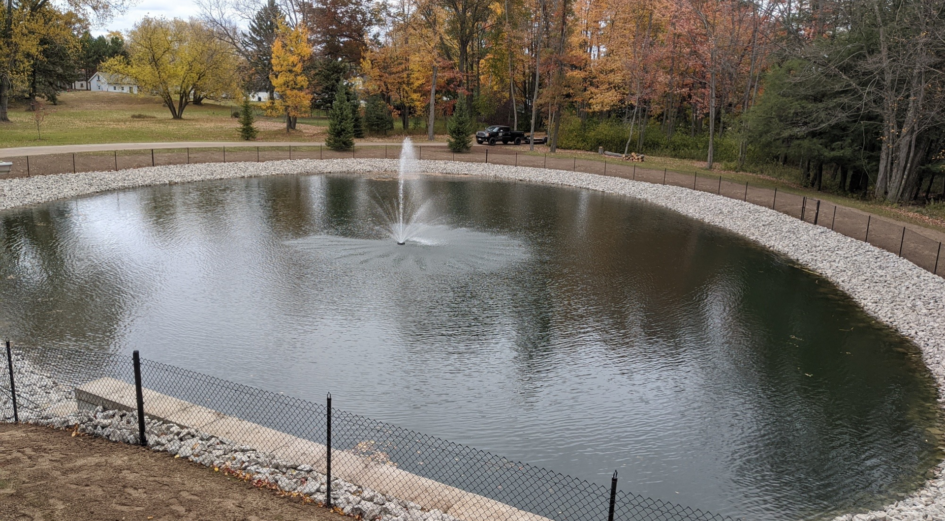 Oval-shaped pond with a fountain in the center. The banks are lined with rocks, and trees surround the pond.