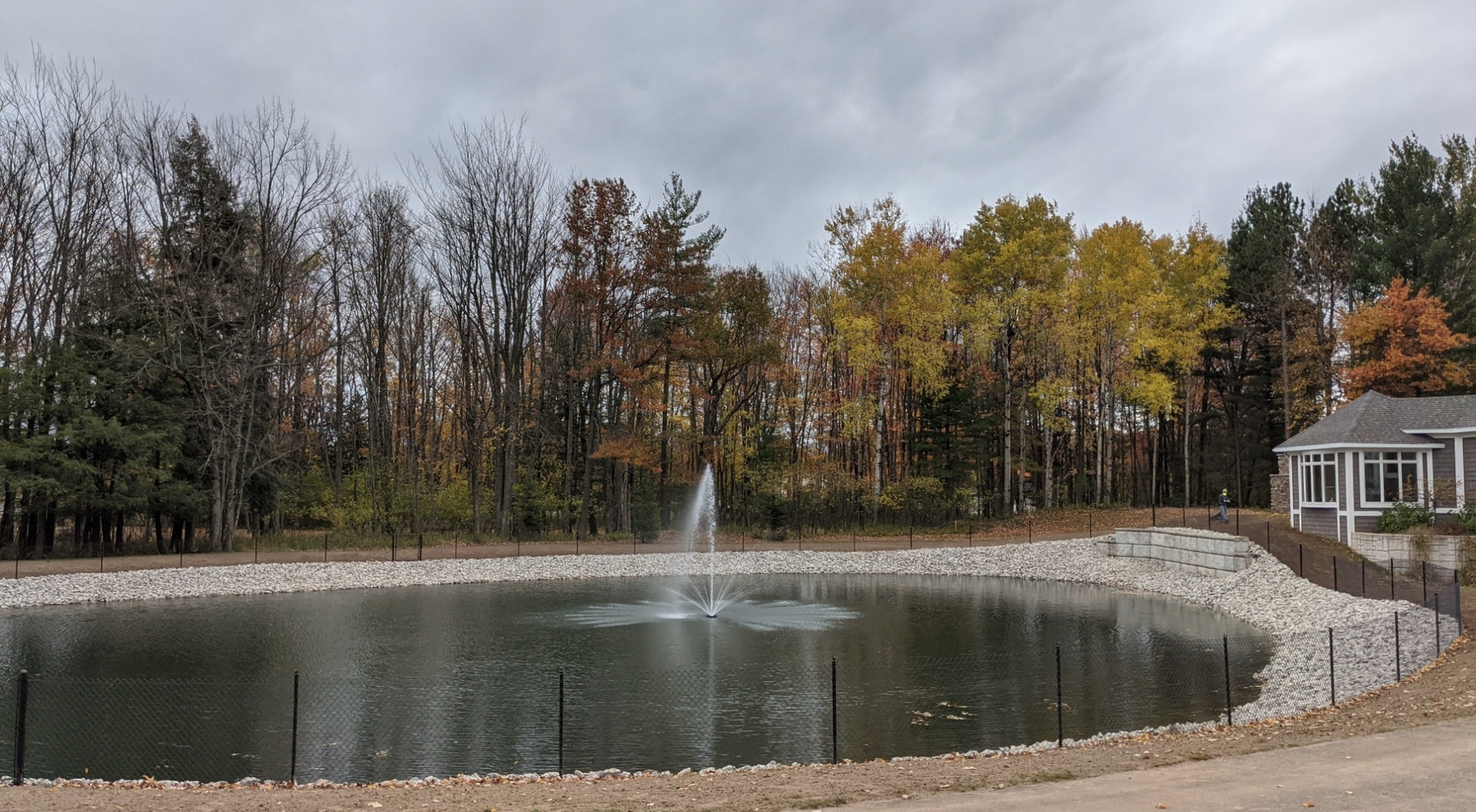 Pond with a fountain, surrounded by trees with fall colors. A white building is visible on the right.