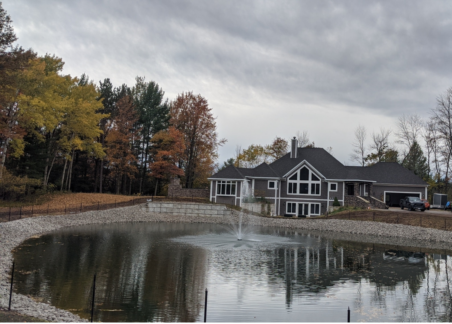 Gray house next to a pond with a fountain, trees, and cloudy sky.