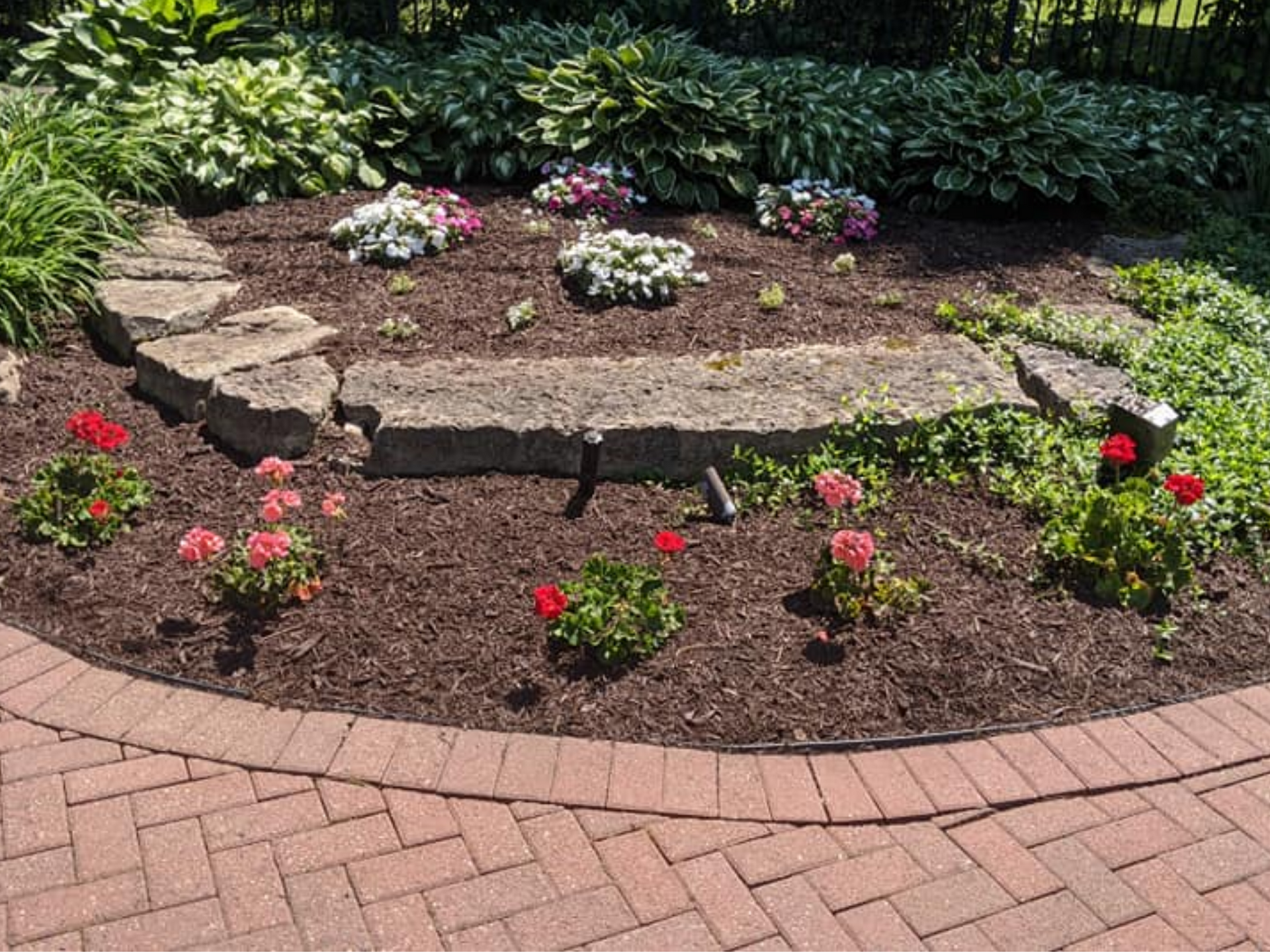 A circular flower bed with red and pink flowers, surrounded by a brick border and mulch.