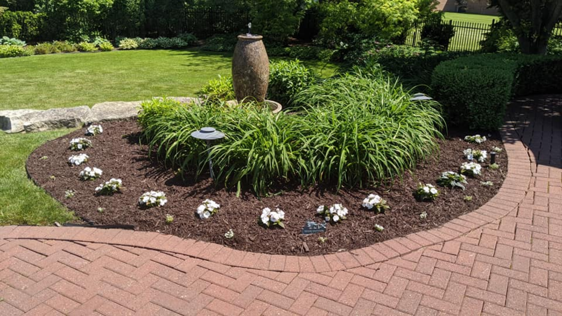 Circular garden bed with fountain, green plants, and white flowers, bordered by brick pathway and lawn.