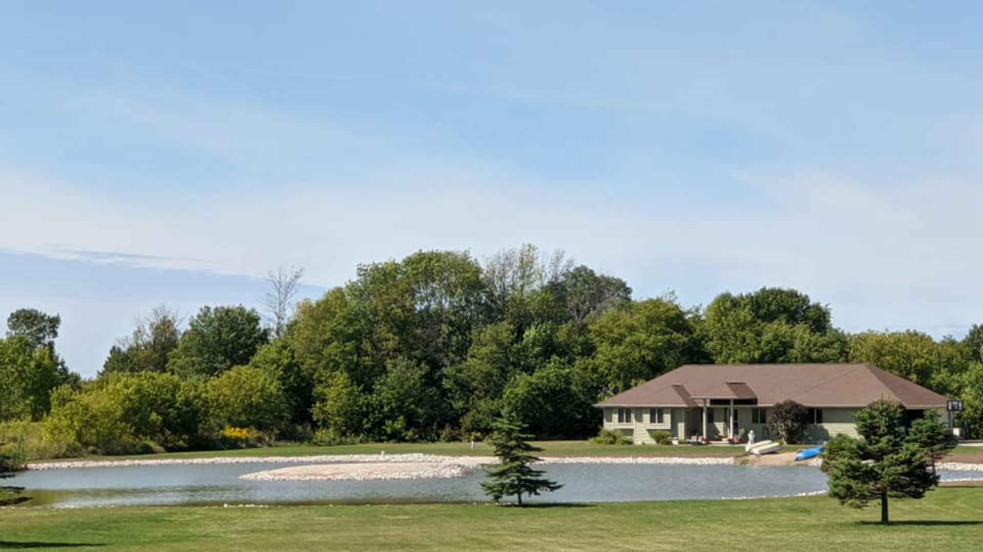 Lakeside house with a brown roof and a backdrop of green trees under a blue sky.