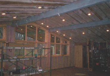 Wooden loft interior with exposed beams, shelves, and warm ceiling lights at night.