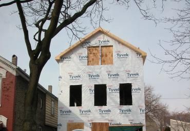 House under construction wrapped in Tyvek, with boarded windows and a large tree in front