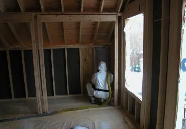 Framed interior of a house under construction, with a worker kneeling by a wall opening near a window.
