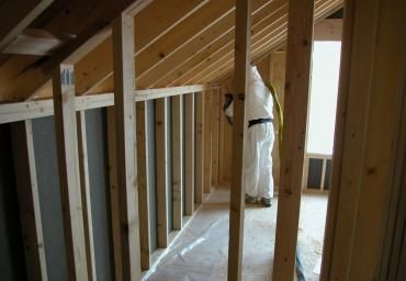 Attic framing with exposed wooden studs and a worker in white protective suit spraying insulation