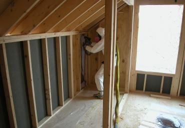 Worker installing insulation in an unfinished attic room with exposed wall framing.