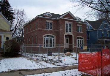 Red brick house under construction in a snowy neighborhood, with orange safety fencing in front.