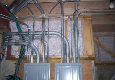 Electrical conduit pipes and circuit breaker panels mounted on an unfinished basement wall.
