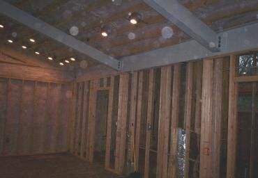 Exposed wooden-framed room with unfinished walls, ceiling beams, and recessed lights inside a house.