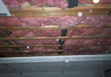 Exposed attic ceiling with pink insulation batts between wooden joists and a gray panel below