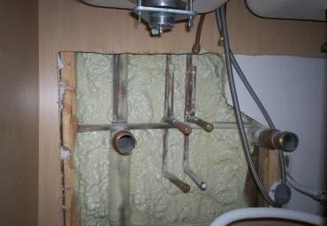 Plumbing pipes and valves protruding from a foam-insulated wall under a sink.