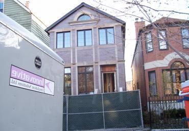 Two-story brick house behind a green fence, flanked by neighboring buildings and a sidewalk sign.