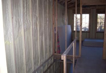 Narrow unfinished hallway with exposed framing, plastic-covered wall, and a blue floor mat by a window.
