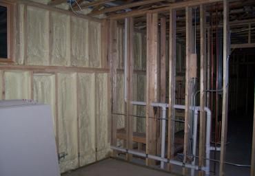 Unfinished basement wall framing with exposed studs, insulation, and drywall sheets stacked on the left.