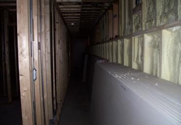 Narrow unfinished hallway with exposed wooden framing and a long white panel leaning along the right wall.