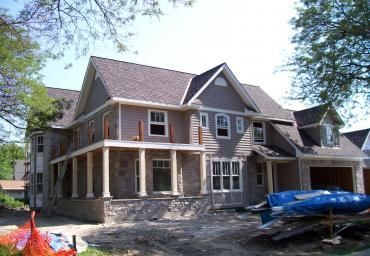 Large gray house under construction with a wraparound porch and blue tarp-covered materials in the yard