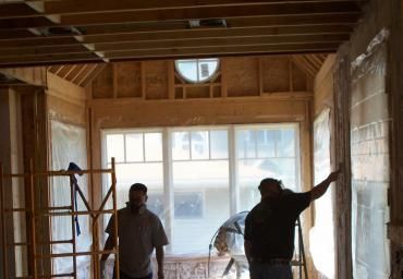 Two people stand in a framed room under construction with ladders and exposed wood beams.