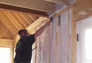 Worker installing insulation in a wooden interior wall beneath exposed ceiling beams.