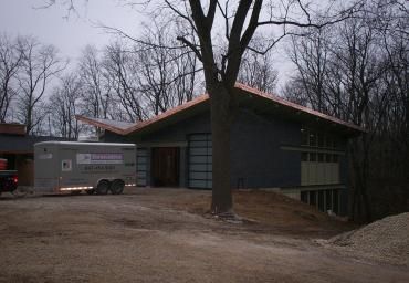 Wooded hillside home with a dark exterior and a trailer parked beside it on a dirt driveway
